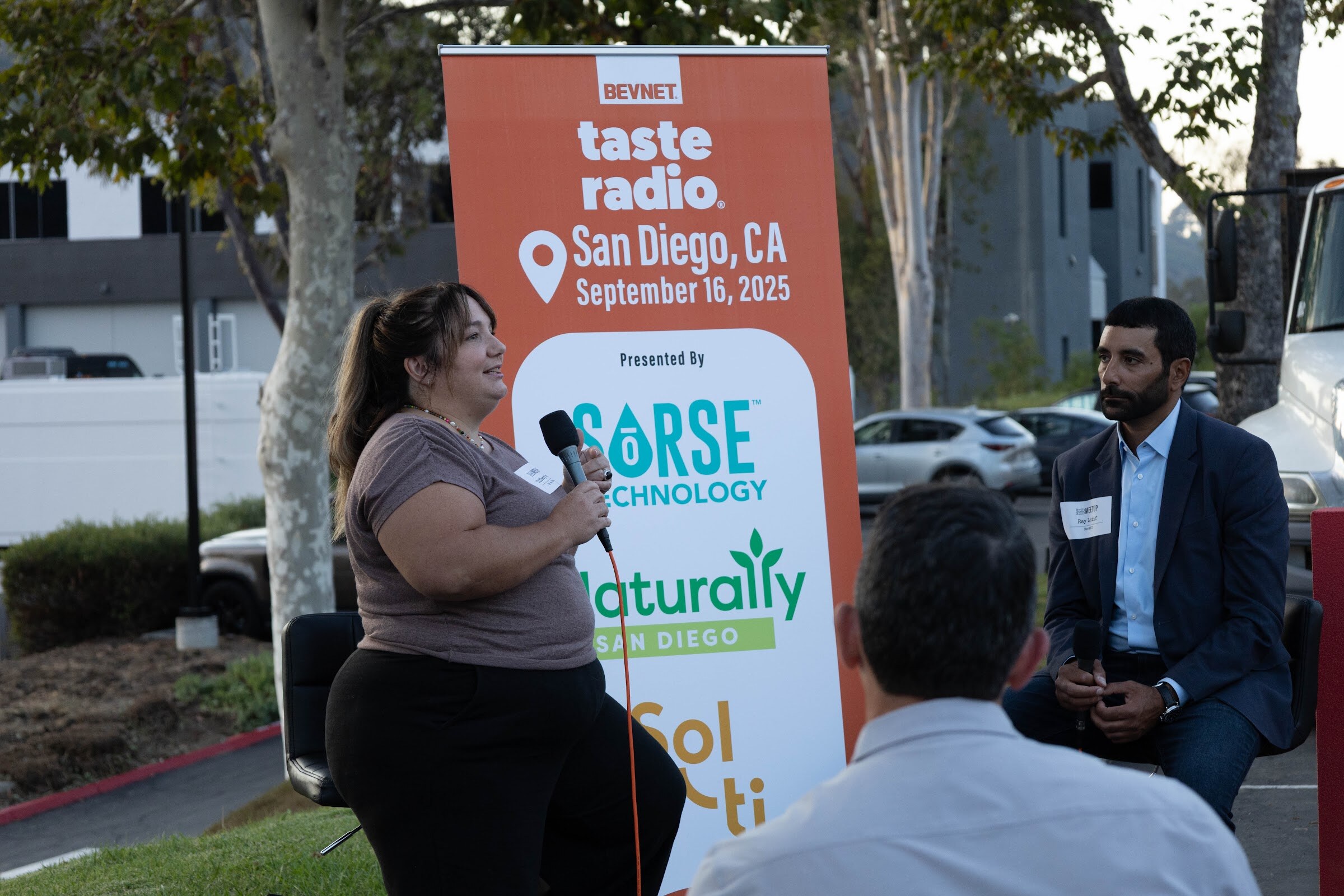 Woman in a gray top holding a microphone in front of the banner, with the host seated at right.