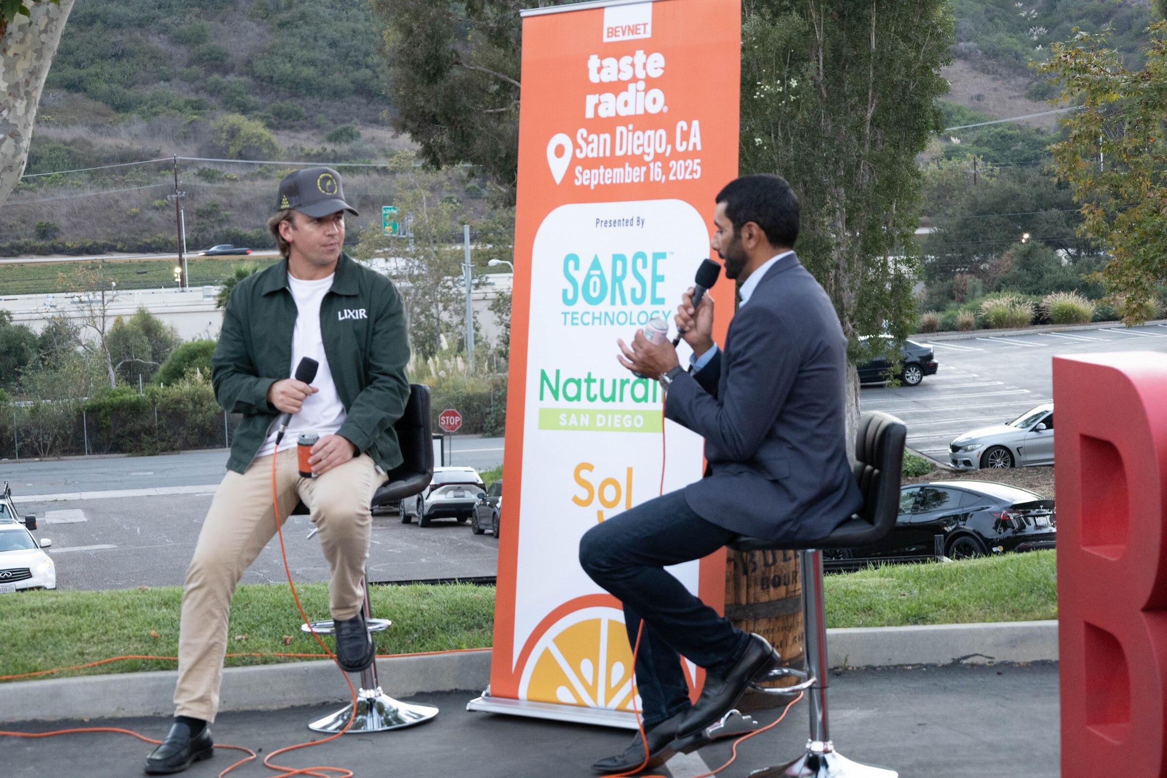 Two men on stools beside the banner, one in a green jacket and cap.