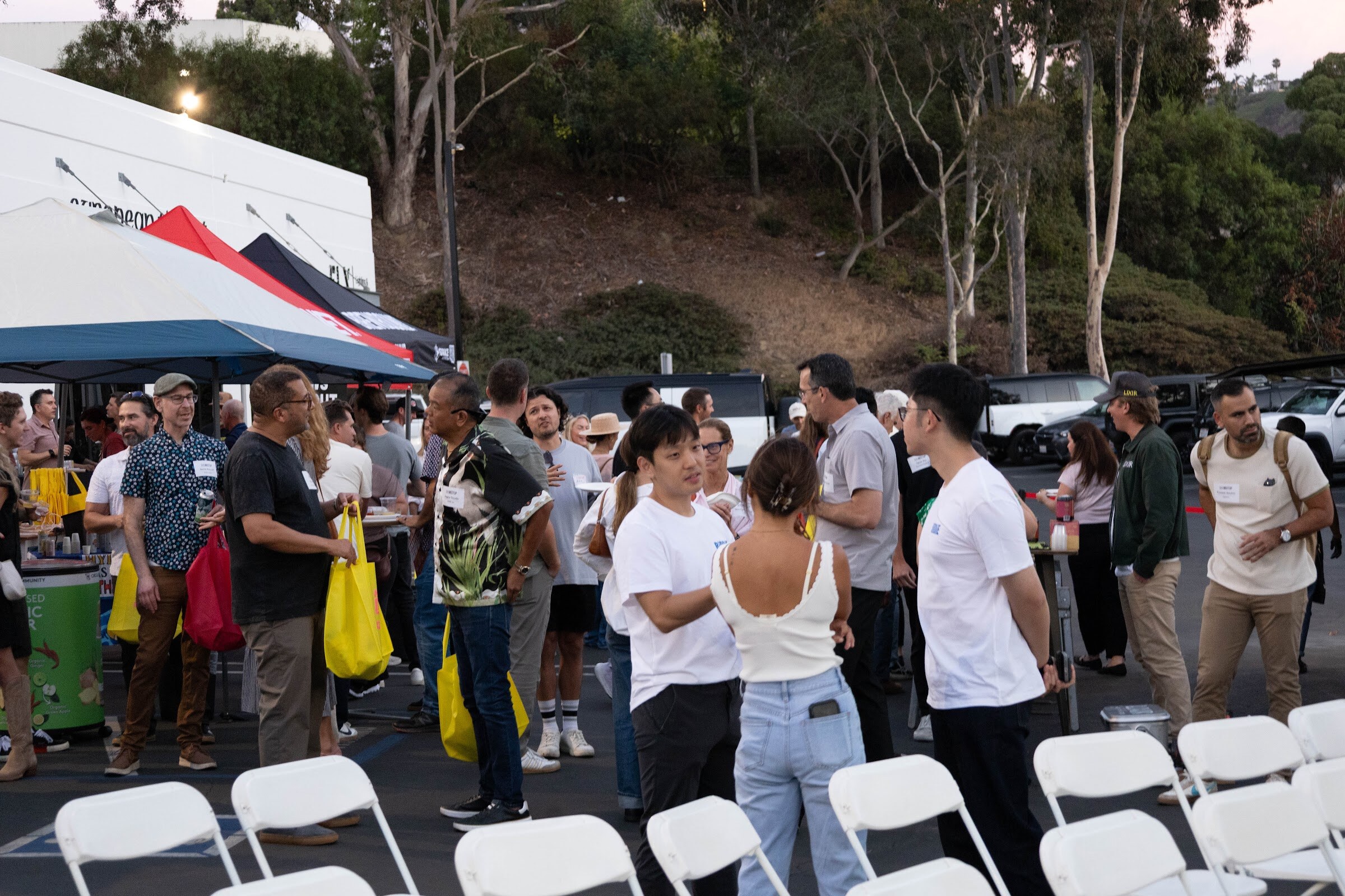 Crowd standing between chairs and tents at dusk, with yellow tote bags.