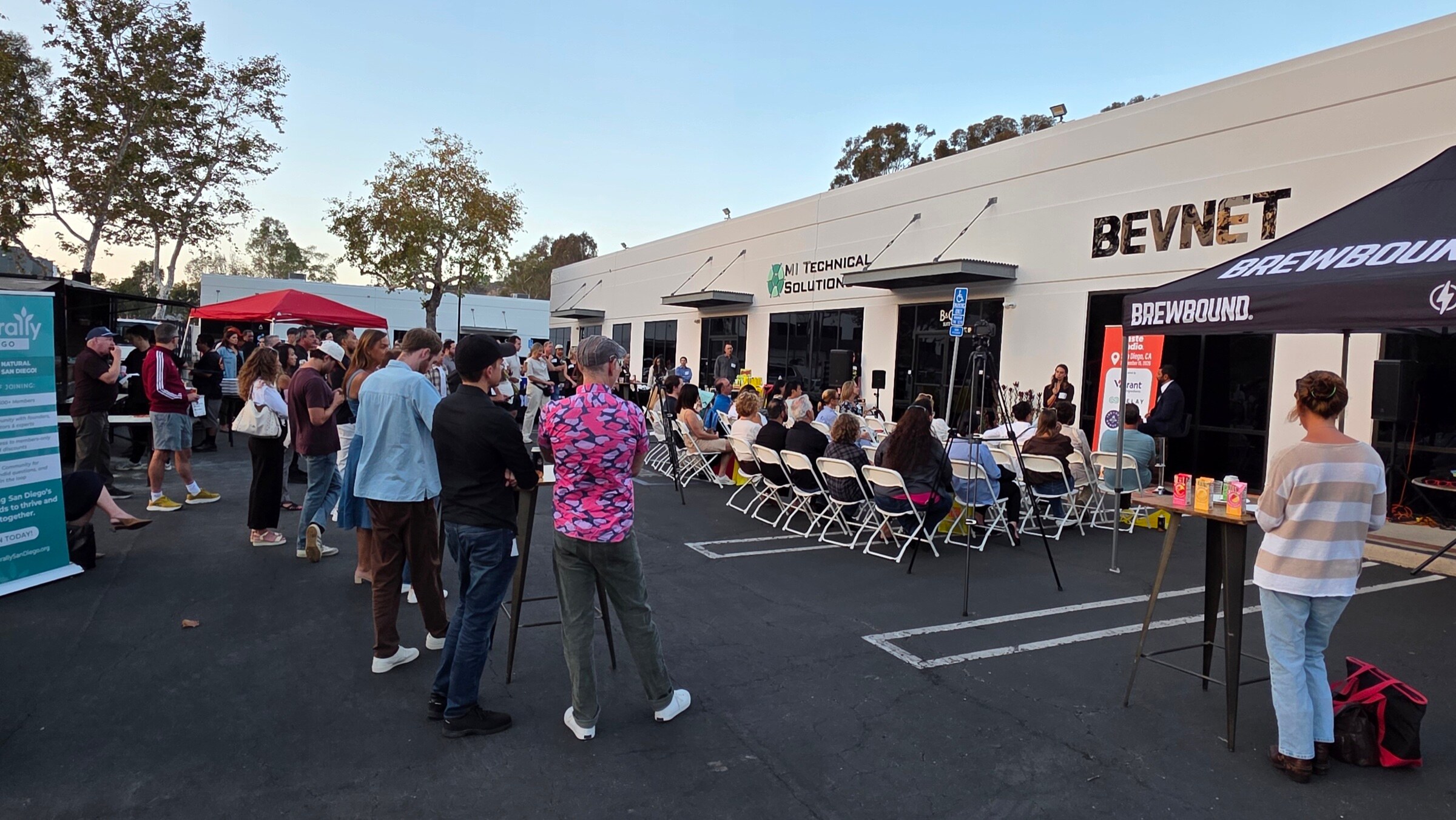 Crowd in a parking lot outside a white building with a BevNET sign, rows of folding chairs facing a Taste Radio banner and a black Brewbound tent at right.