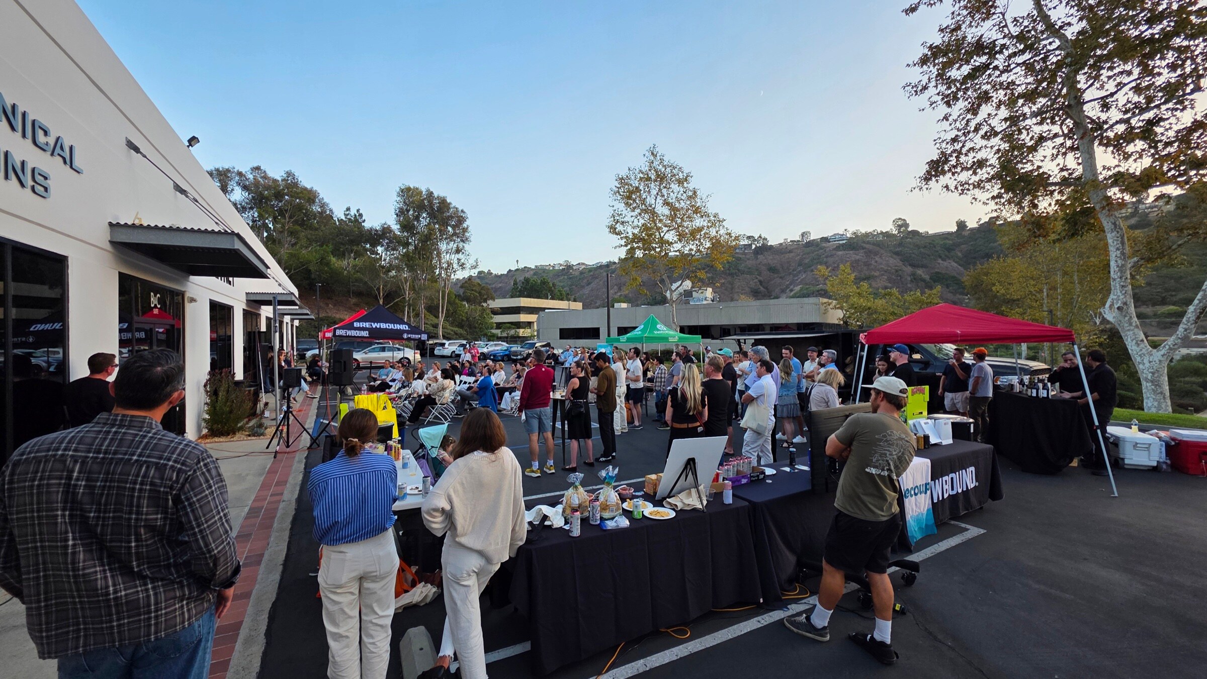 Wide view of attendees between sampling tables in a parking lot, with Brewbound, green Naturally San Diego and red event tents and hills behind.