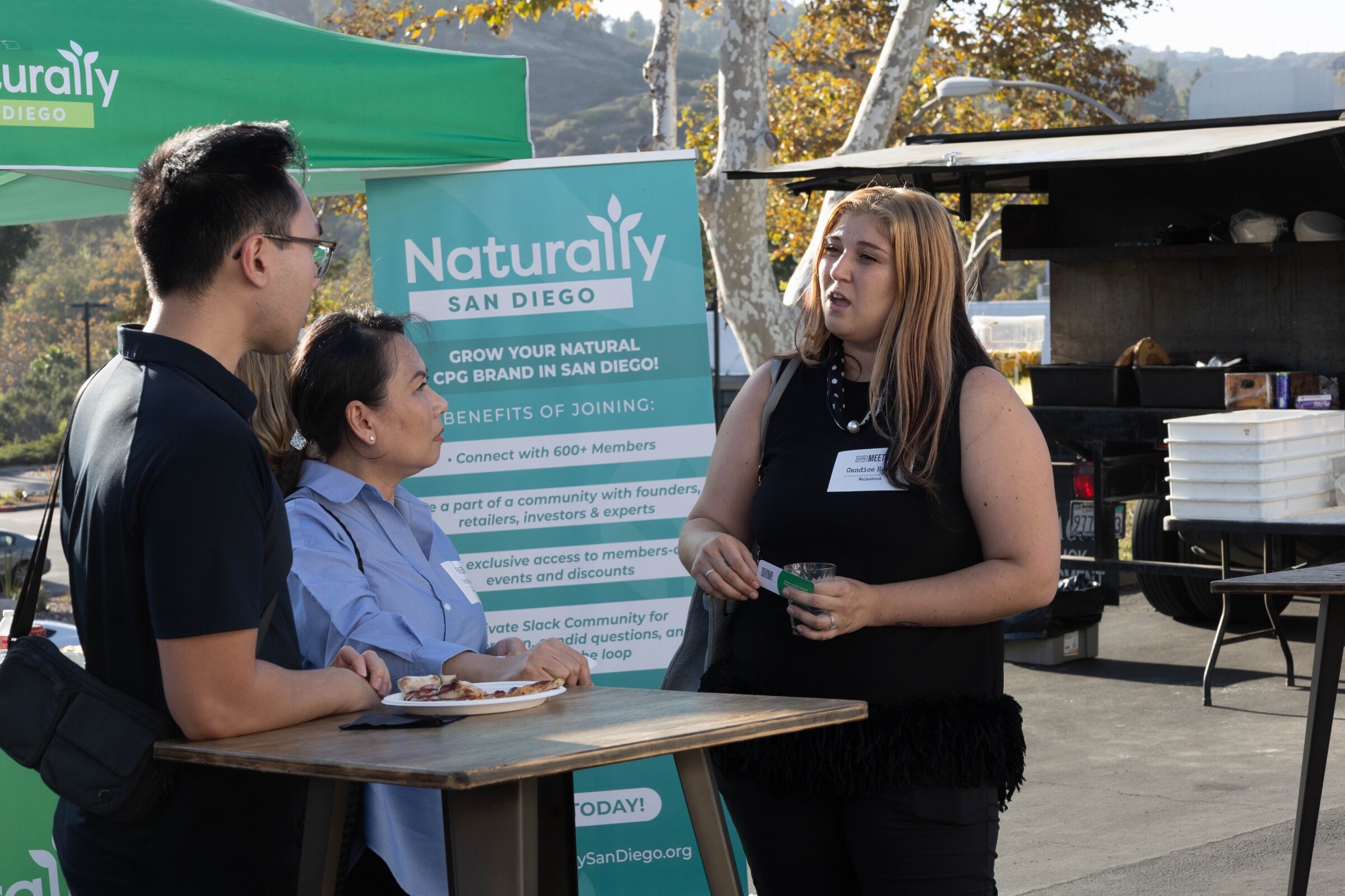 Three attendees talk at a high-top table beside a teal Naturally San Diego banner listing the benefits of joining.