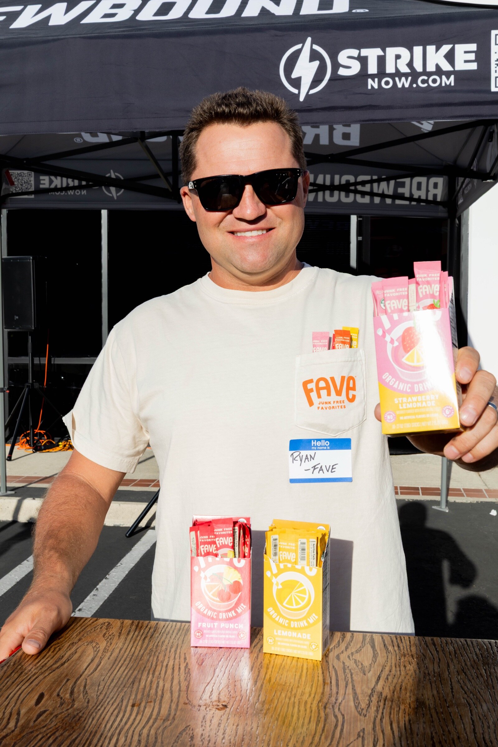Man in a Fave T-shirt and sunglasses holds up a box of Fave Strawberry Lemonade organic drink mix behind boxes of Fruit Punch and Lemonade.