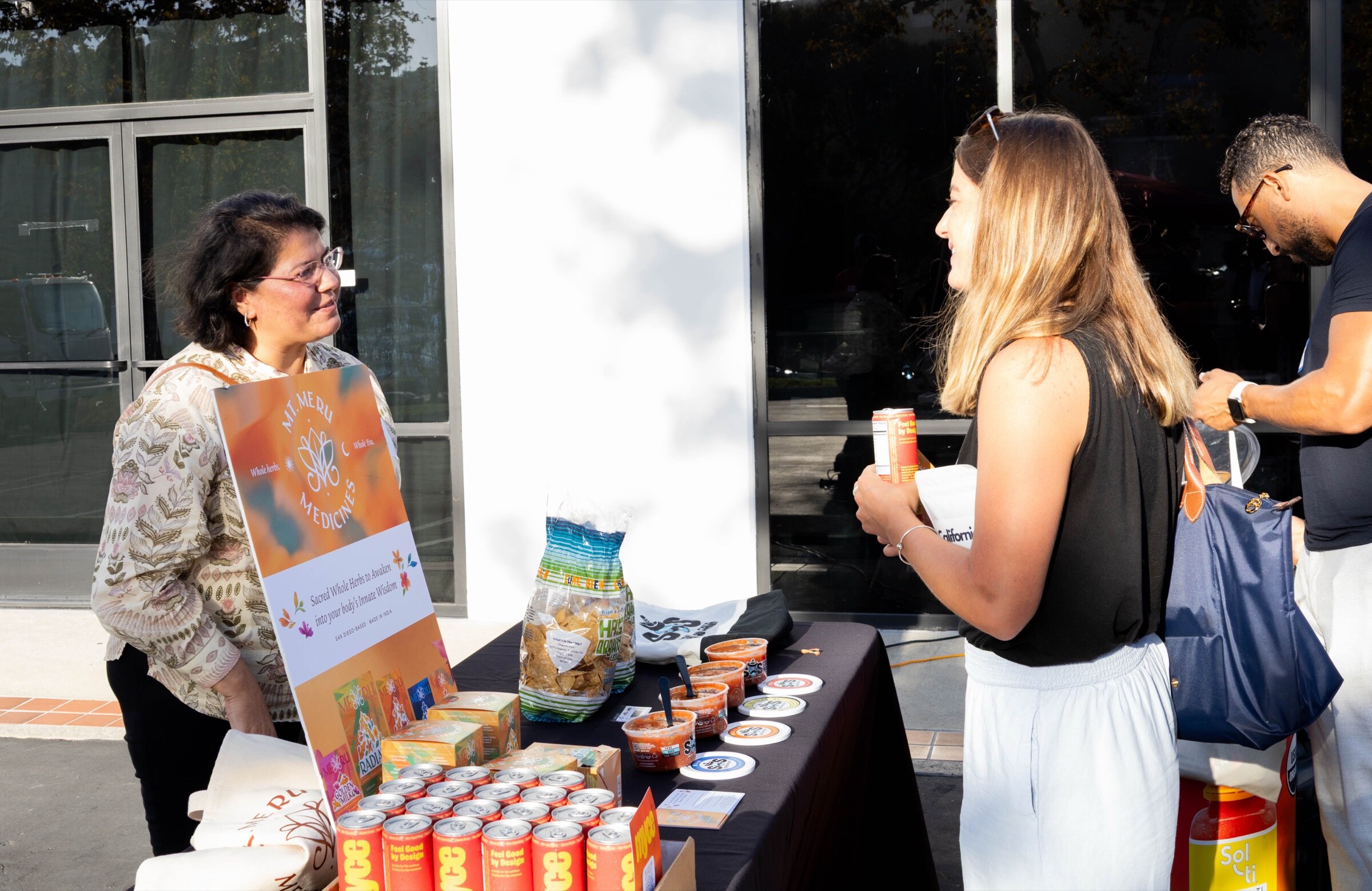 Woman behind a Mt. Meru Medicines sign talks with an attendee at a table of Myce cans, salsa tubs and chips.