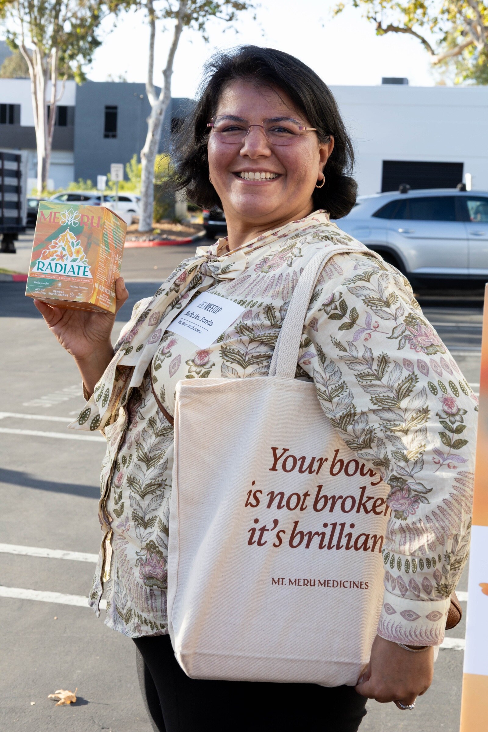 Smiling woman carrying a Mt. Meru Medicines tote holds up a box of Radiate herbal tea.