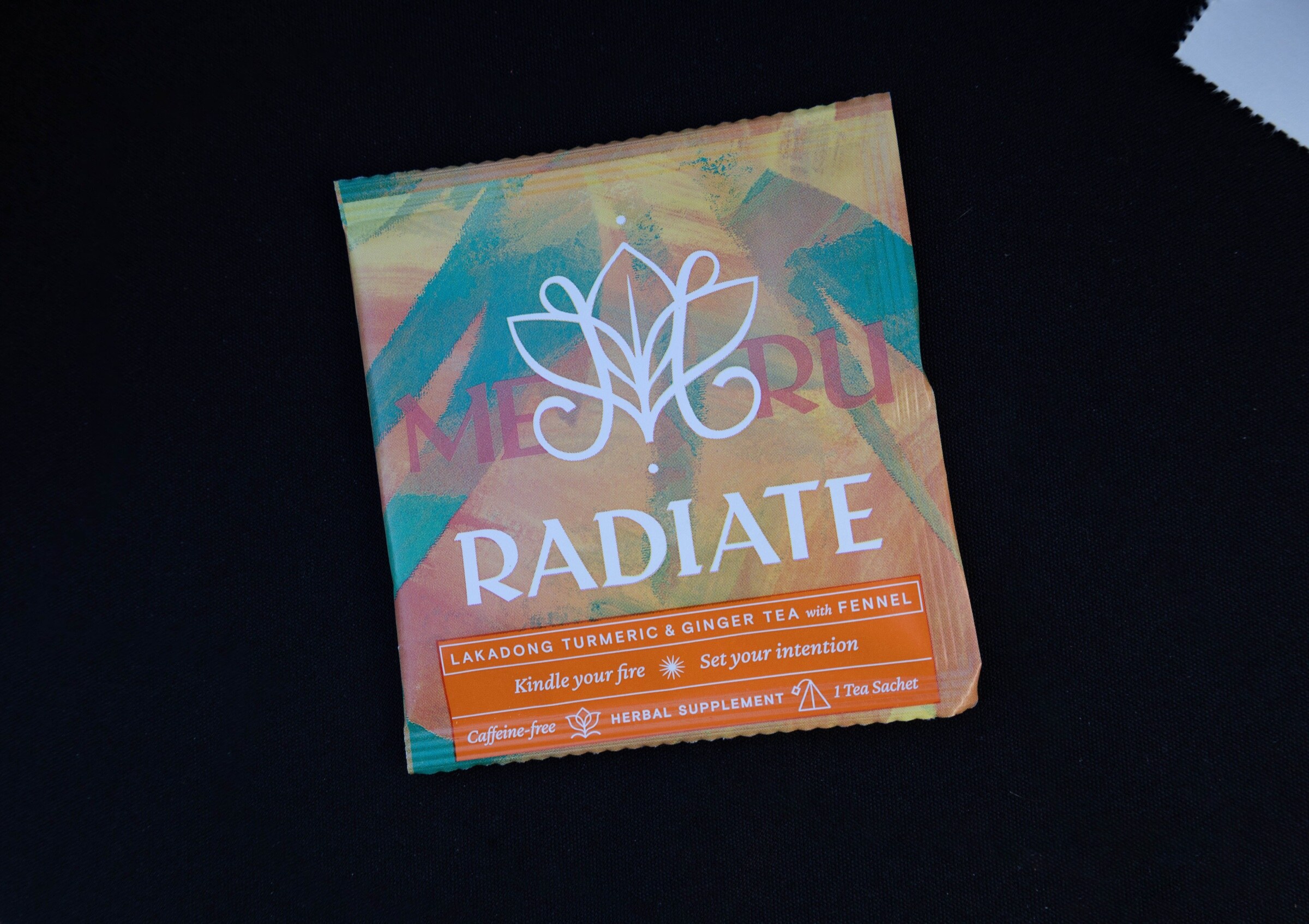 A single Radiate herbal tea sachet with an orange and teal label on a black tablecloth.