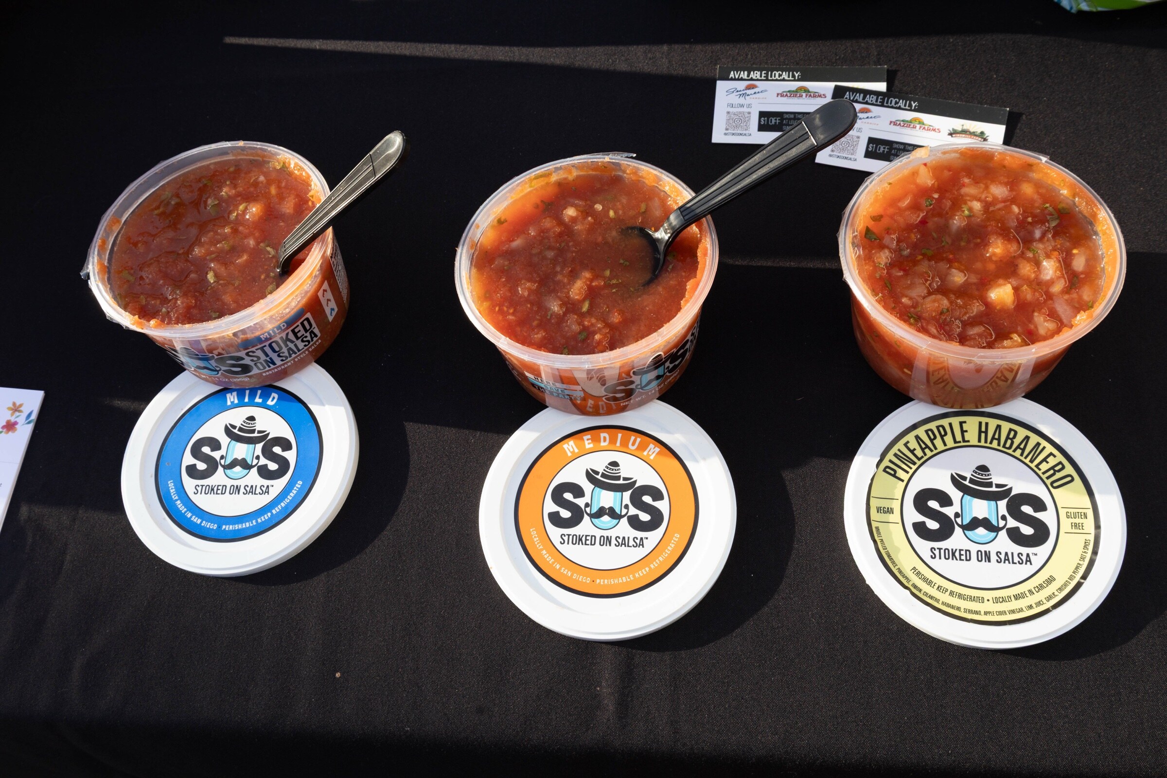 Three open tubs of Stoked on Salsa with serving spoons, their lids labeled Mild, Medium and Pineapple Habanero.