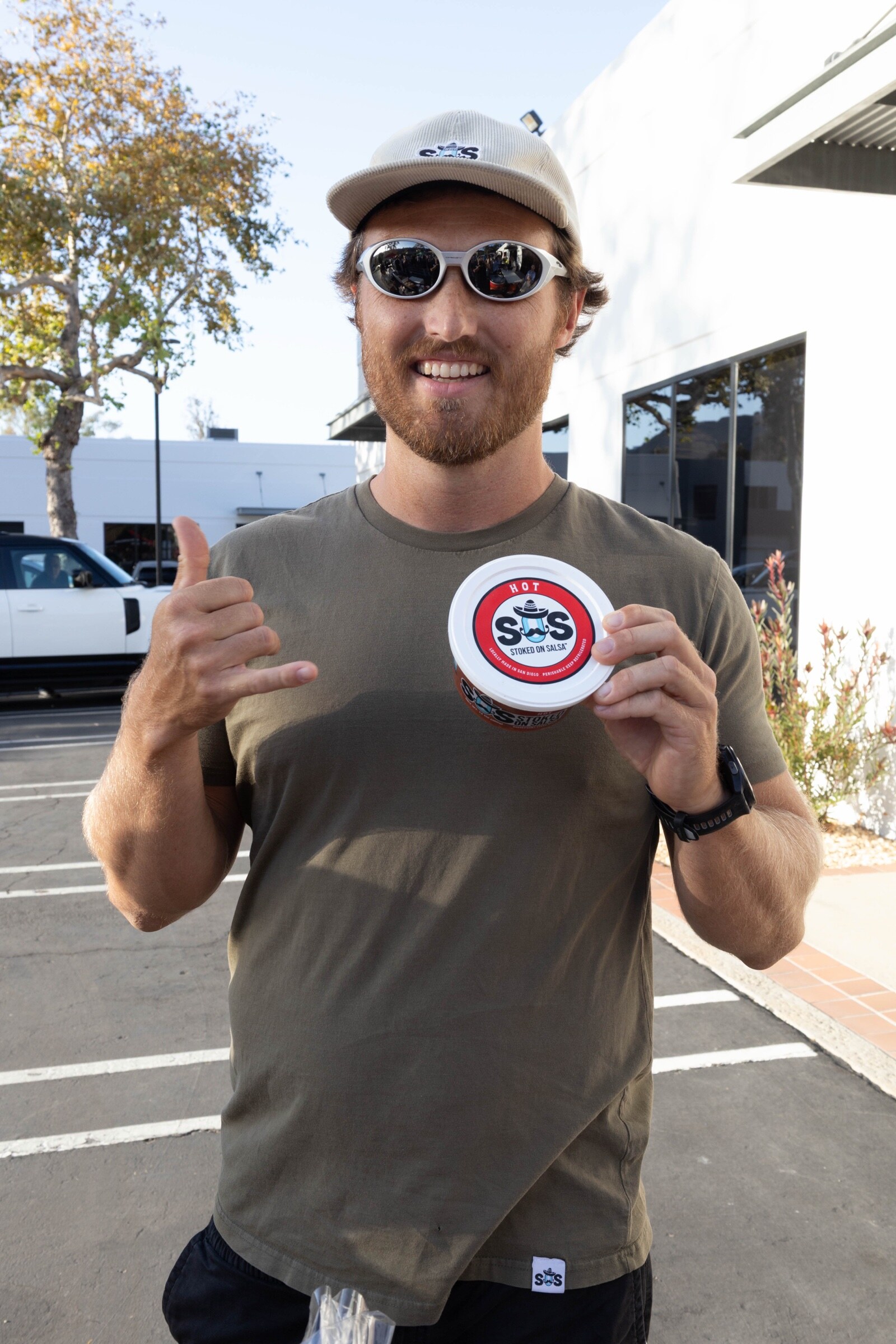 Smiling man in a Stoked on Salsa cap and mirrored sunglasses makes a shaka sign while holding a tub of Hot salsa.