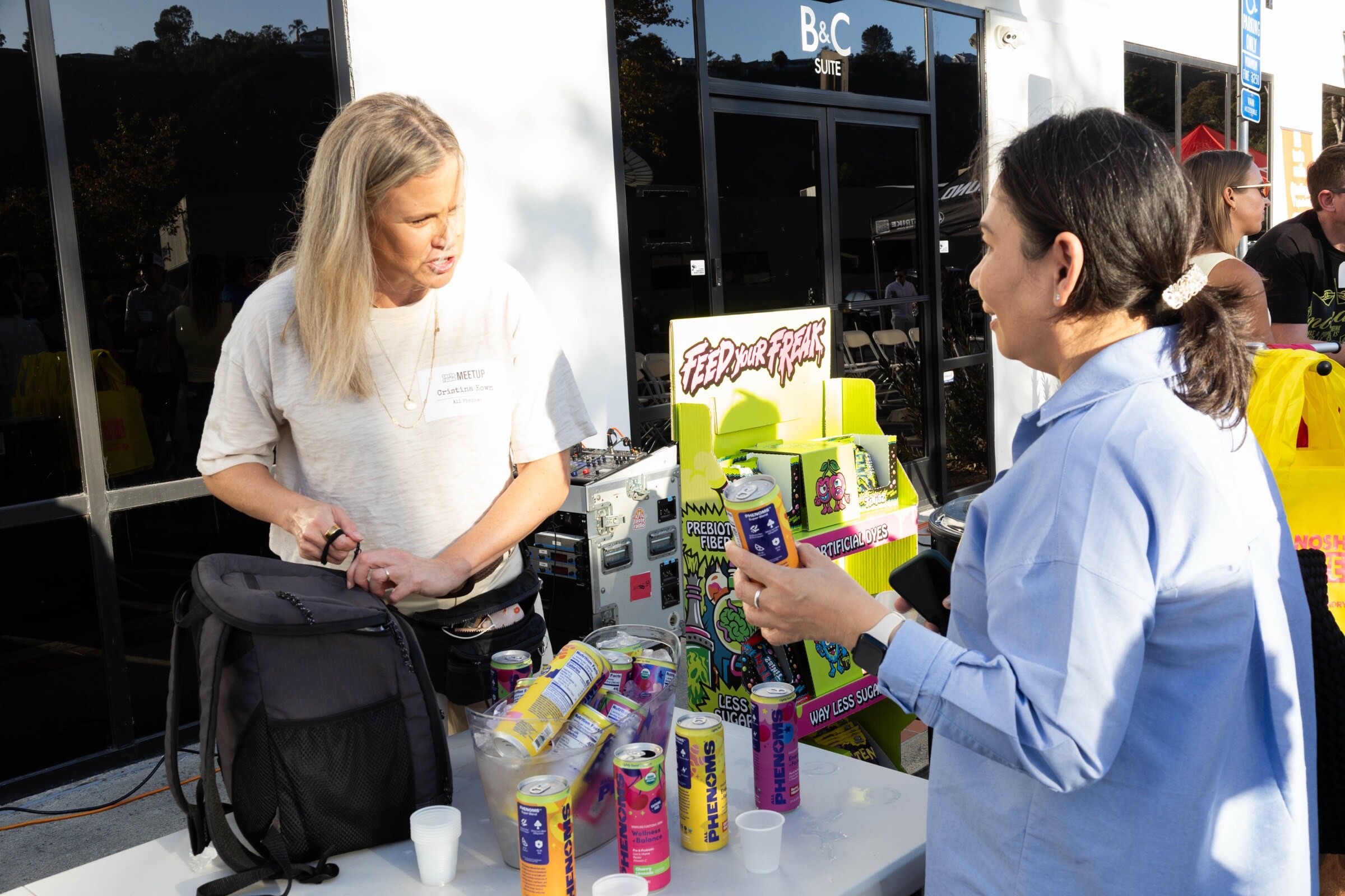 Woman reaches into a bag beside a tub of Phenoms cans and a yellow Rotten display while an attendee holds a can.