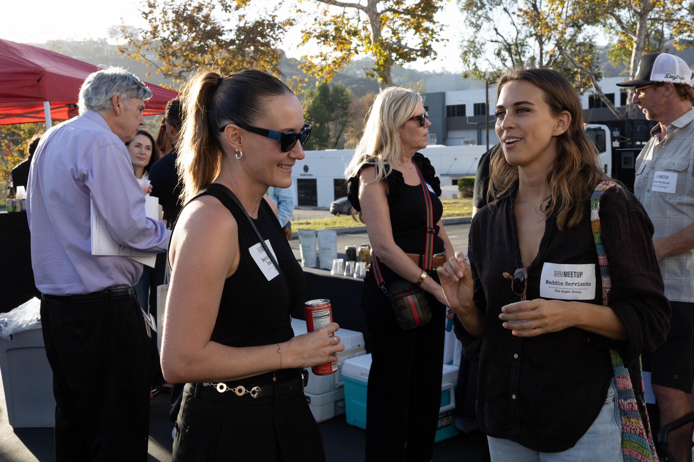 Two women talk and smile, one holding a can, with other attendees and a red tent behind them.