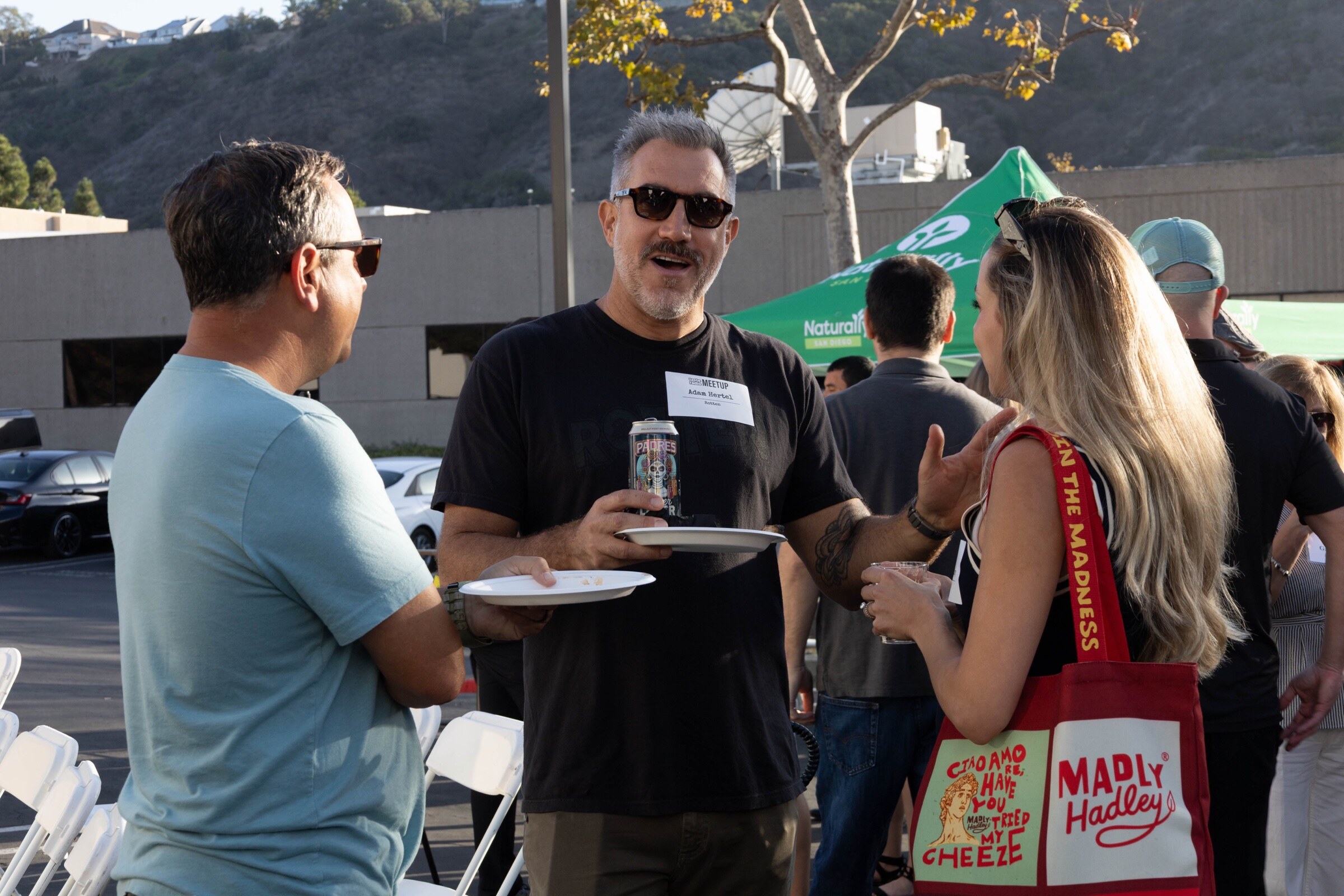 Man in sunglasses holding a plate and a can talks with two attendees, one carrying a Madly Hadley tote bag.