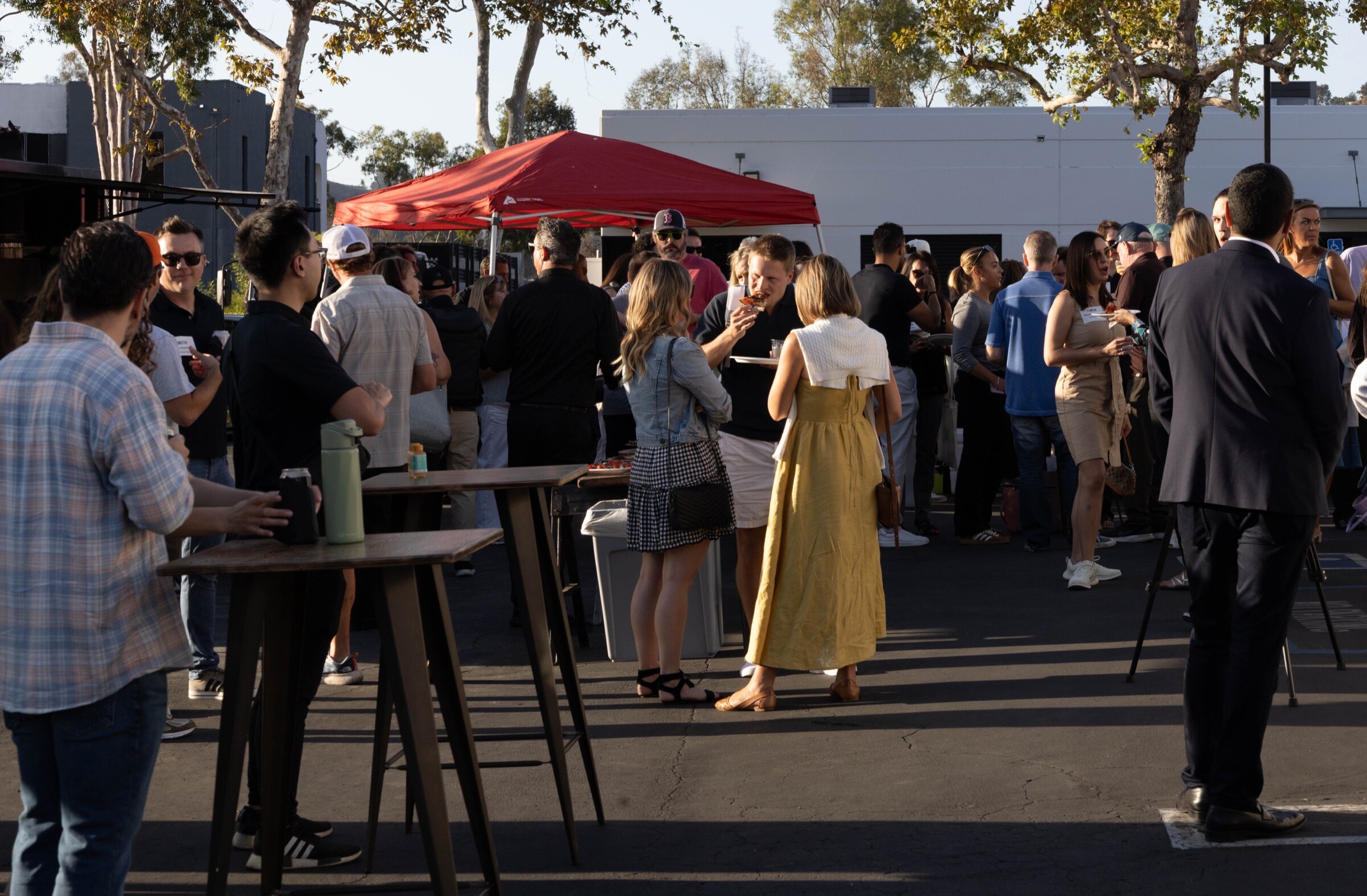 Attendees stand in groups around high-top tables in a sunlit parking lot, with a red tent and trees behind.