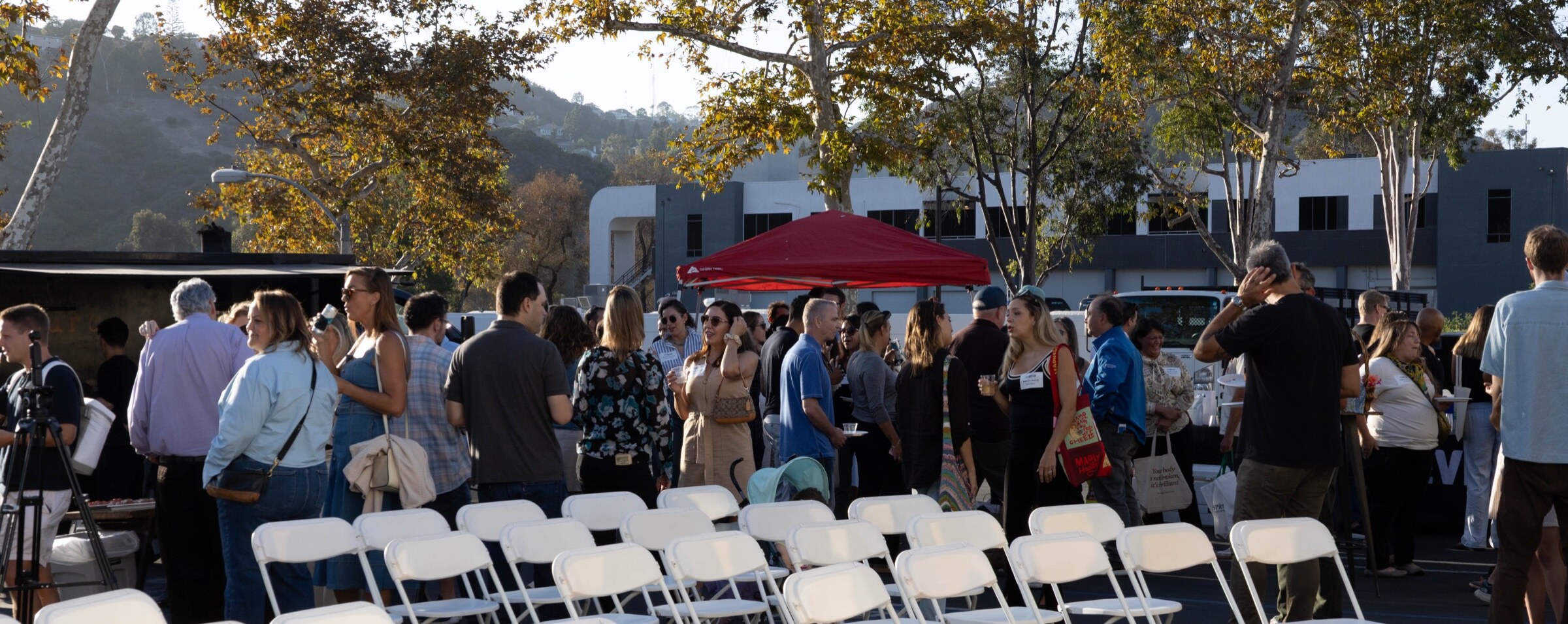 Panoramic view of attendees mingling behind rows of empty white folding chairs, with a red tent and autumn trees.