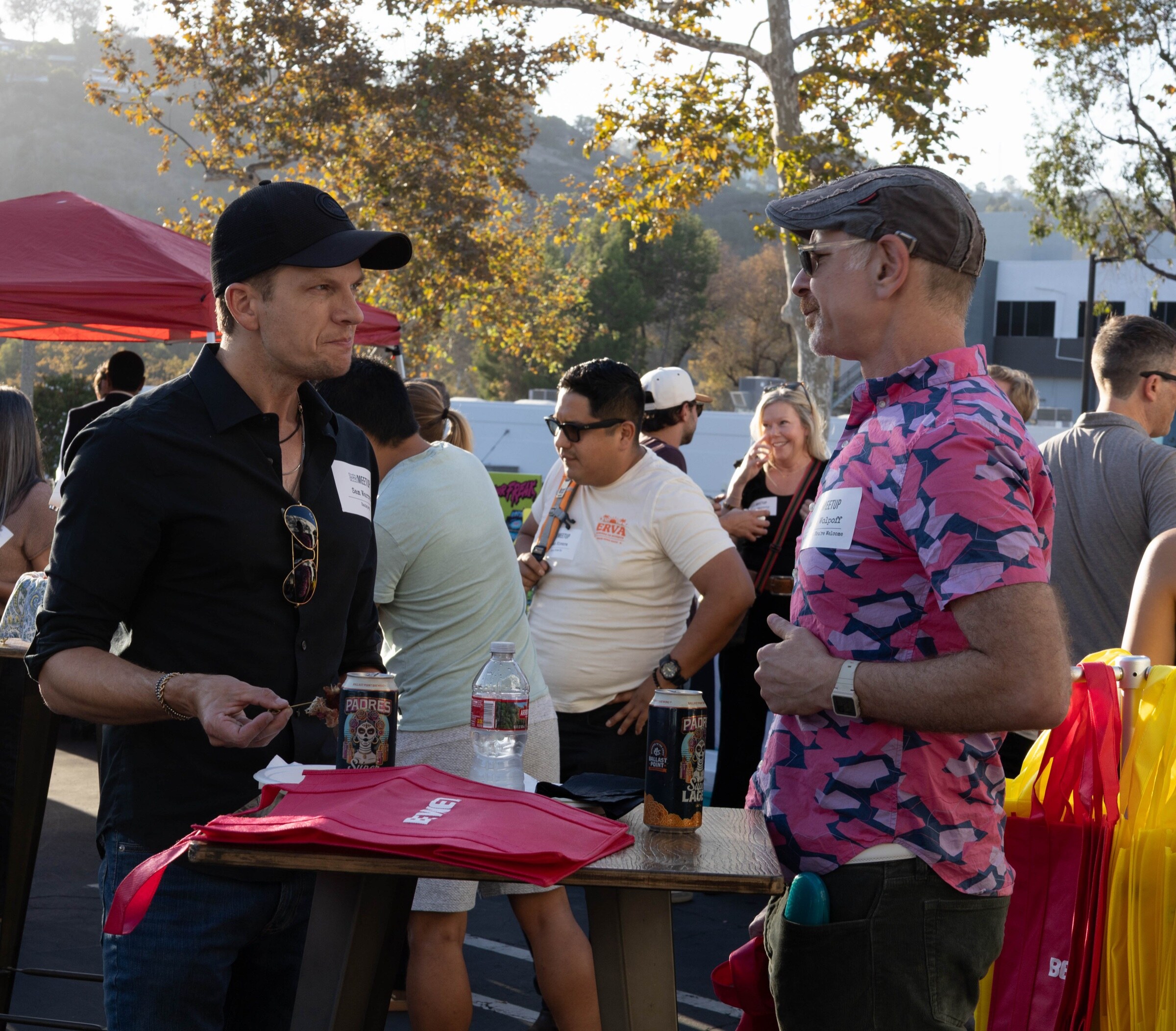 Man in a black cap talks with a man in a pink patterned shirt at a high-top table holding red BevNET tote bags and Padres beer cans.