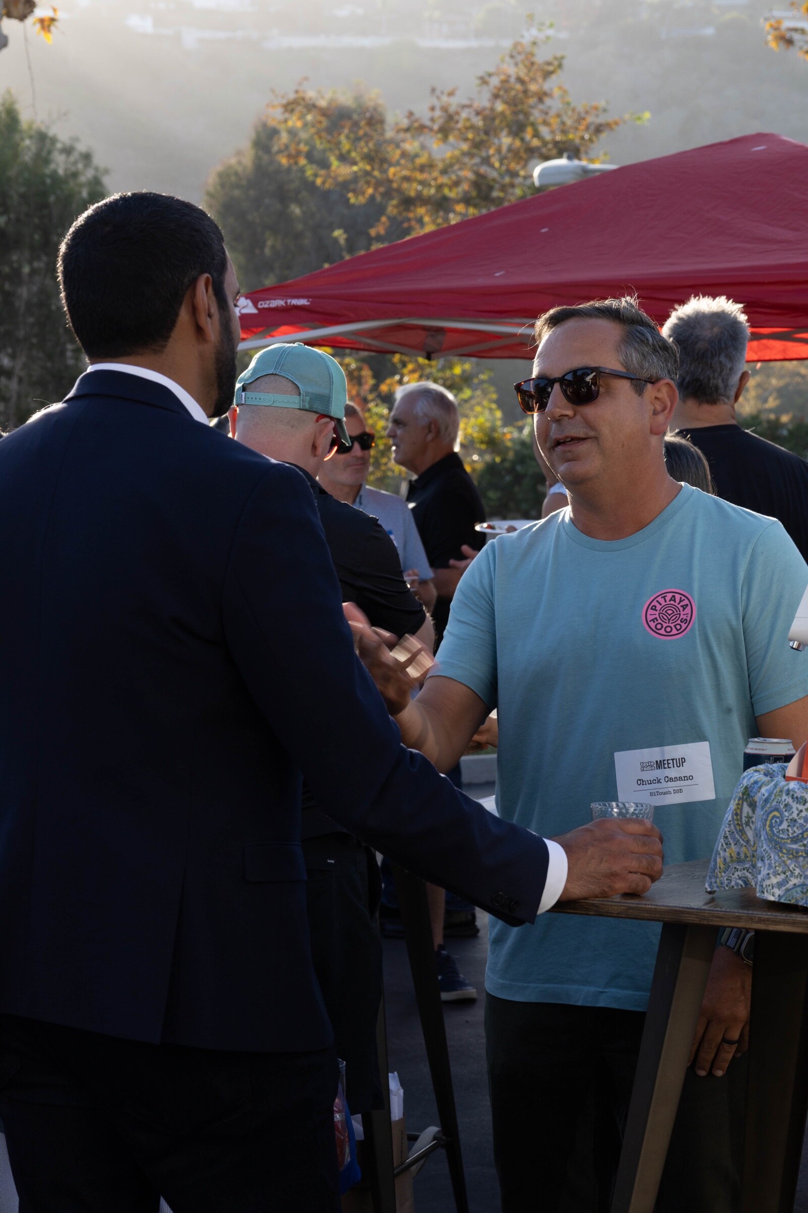 Man in a Pitaya Foods T-shirt and sunglasses talks with a man in a navy suit at a high-top table, backlit by the low sun.