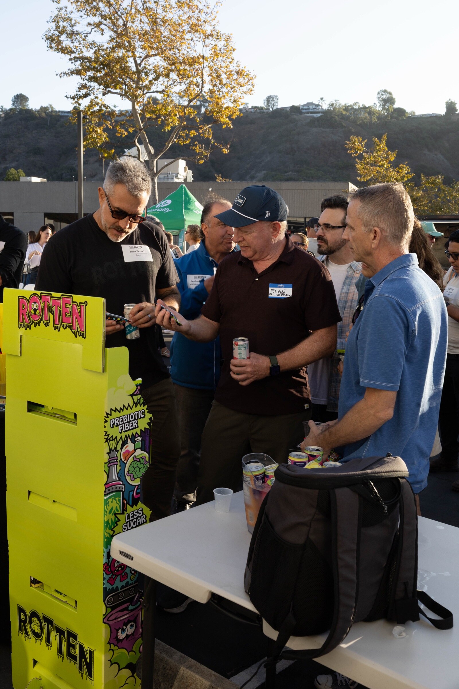 Four men holding cans gather at a table beside a bright yellow Rotten display advertising prebiotic fiber and less sugar.