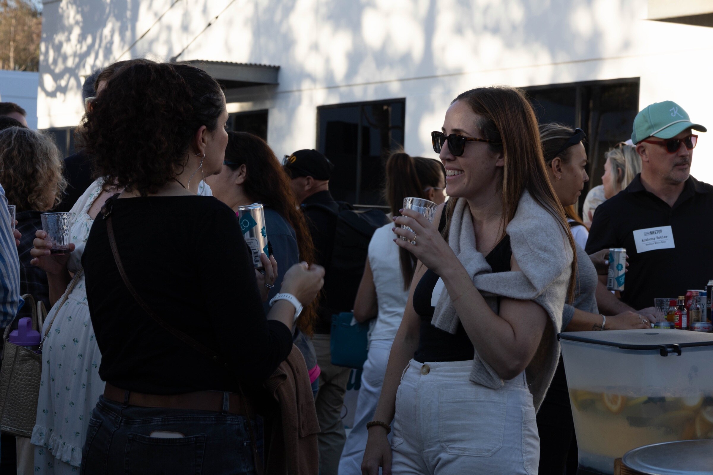 Smiling woman in sunglasses holds a glass while talking with another attendee, with more guests behind.