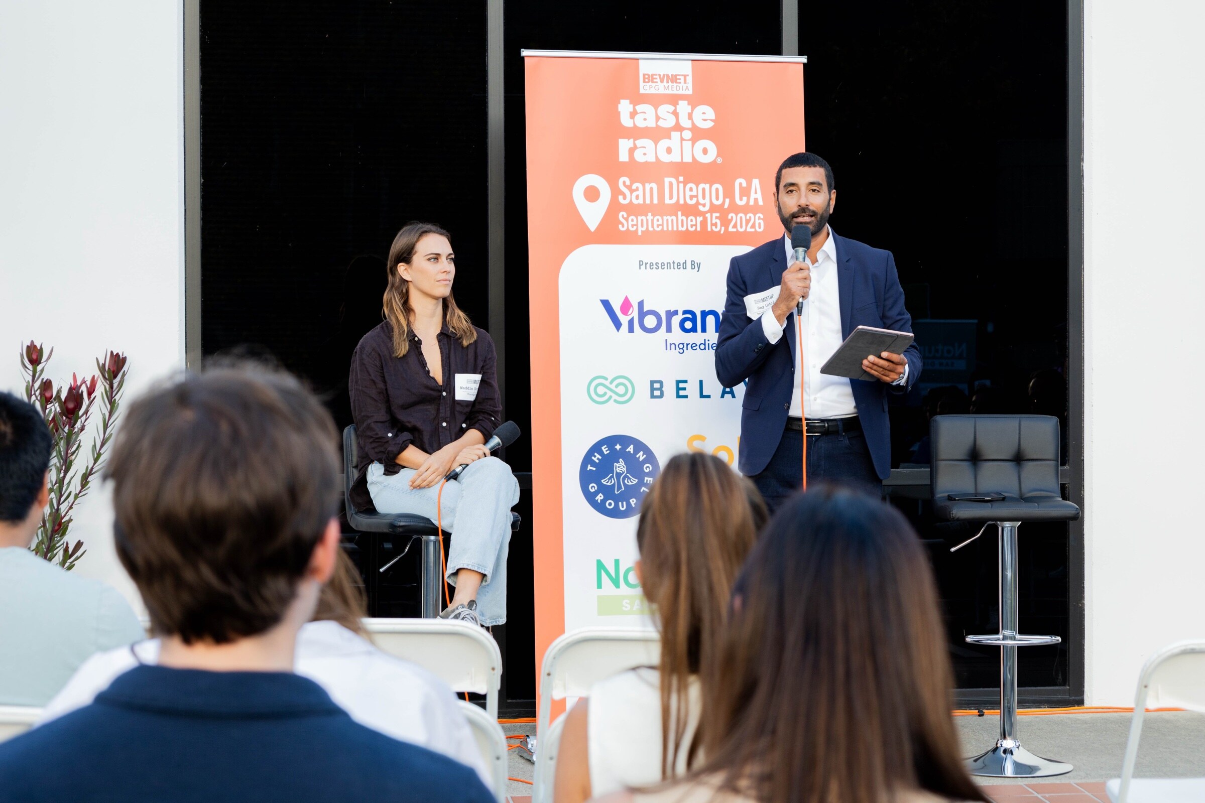 Man in a navy suit speaks into a microphone beside a seated guest and an orange Taste Radio San Diego September 15, 2026 banner, with the audience in the foreground.