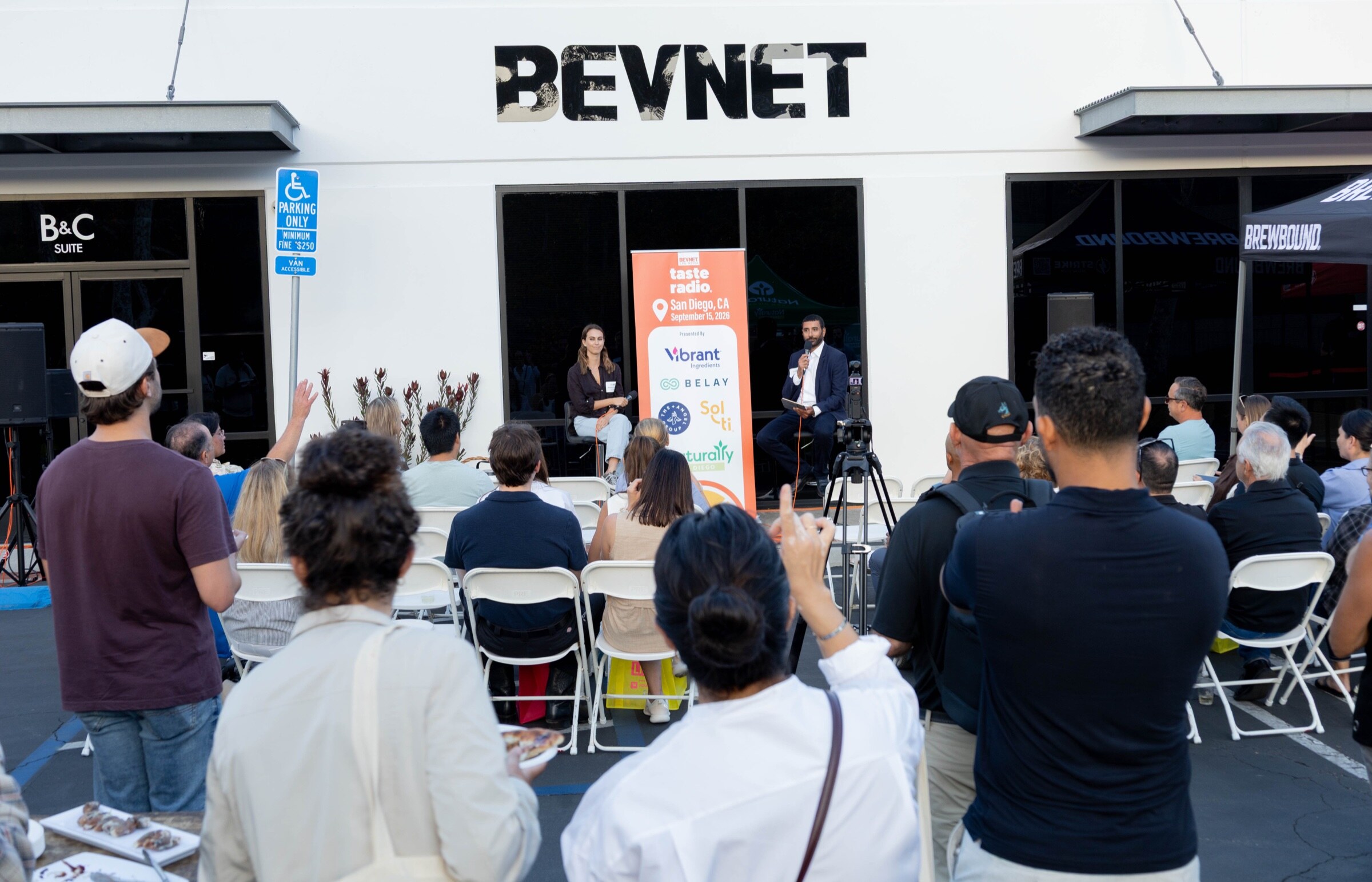 Audience seated and standing in front of a white building with a large BevNET sign, where two people sit on stools beside the Taste Radio banner.