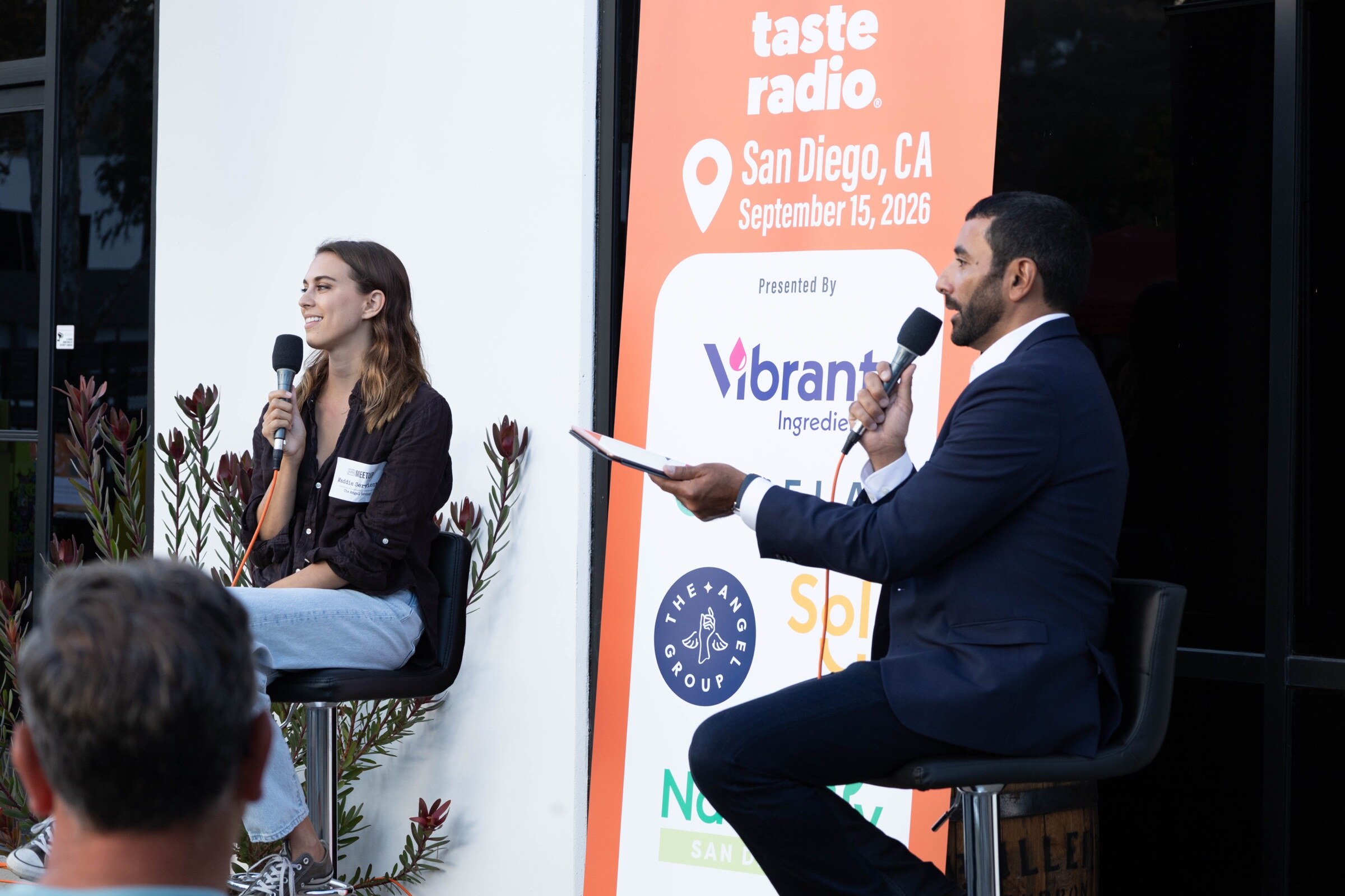 Woman with a microphone smiles on a stool while a man in a navy suit asks a question beside the Taste Radio banner.