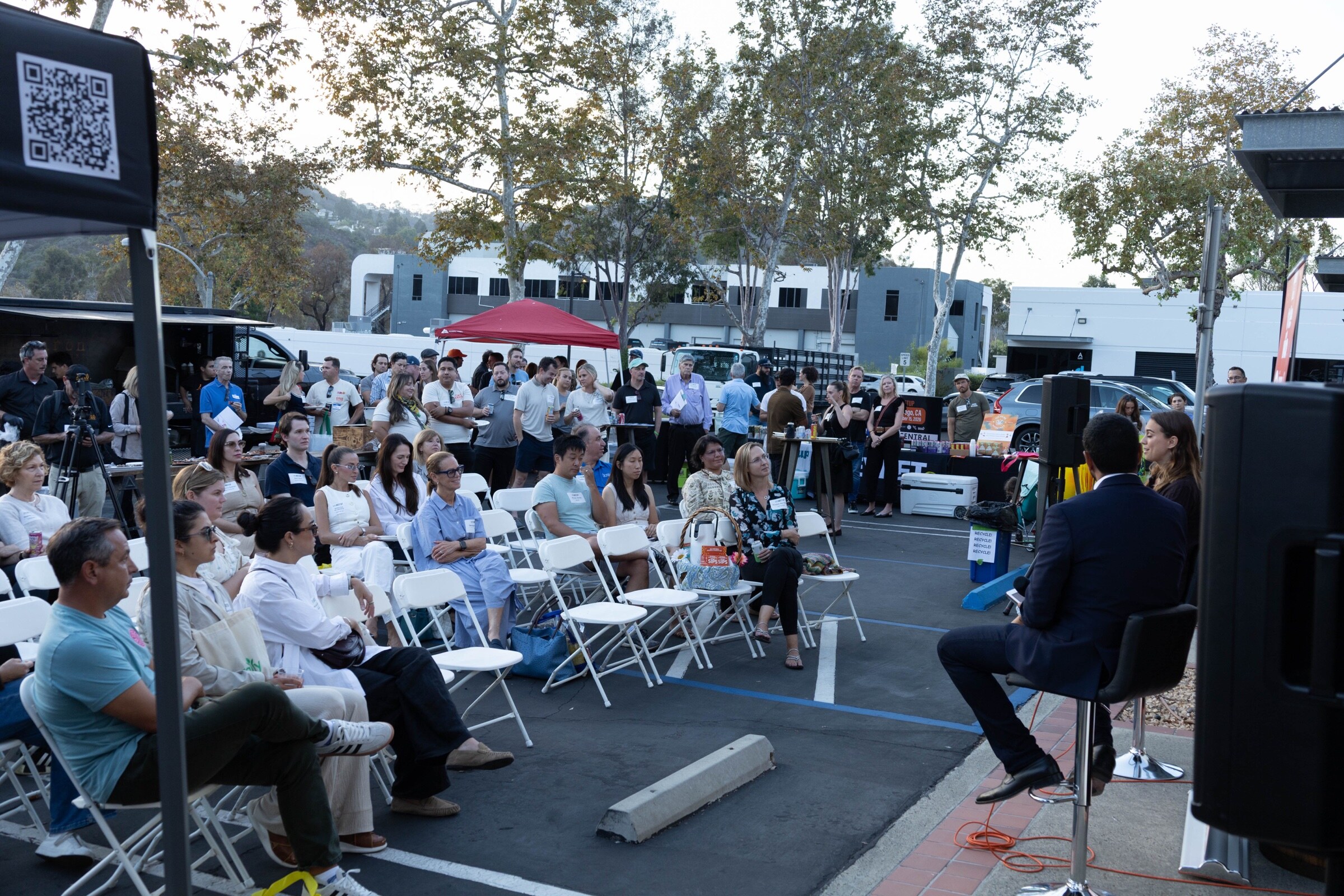 Rows of seated attendees face the stage from the side, with standing guests, a red tent and trees in the background.