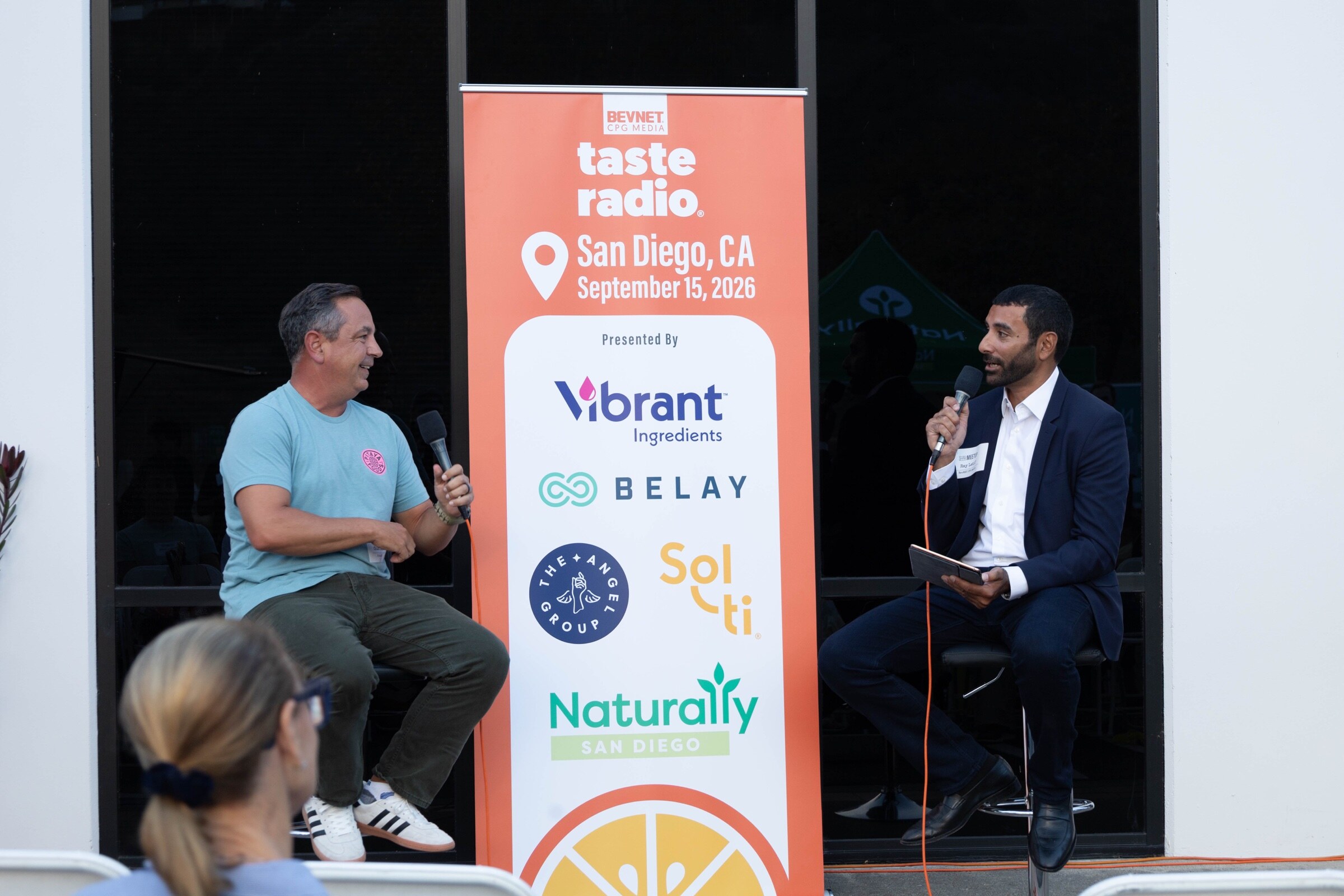 Man in a light blue Pitaya Foods T-shirt talks into a microphone across from a man in a navy suit, with the Taste Radio sponsor banner between them.