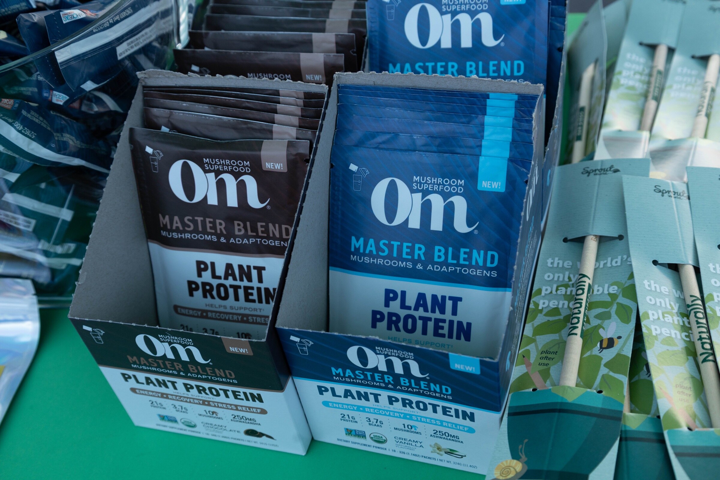 Display boxes of Om Mushroom Superfood Master Blend plant protein packets on a green tablecloth.